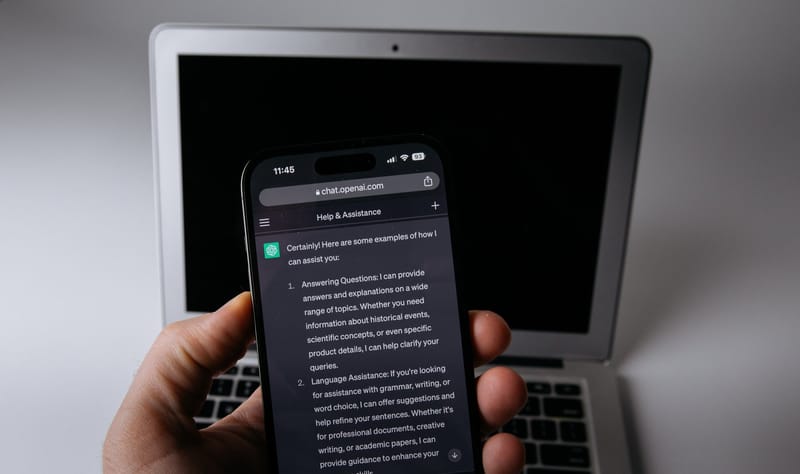 Pessoa segurando um smartphone com uma conversa aberta no aplicativo do ChatGPT, em frente a um notebook